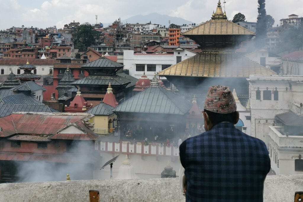 Ein Mann mit traditioneller nepalesischer Kopfbedeckung blickt nachdenklich auf den Tempelkomplex von Pashupatinath in Kathmandu, im Hintergrund Häuser und Rauch vom Tempelritual.