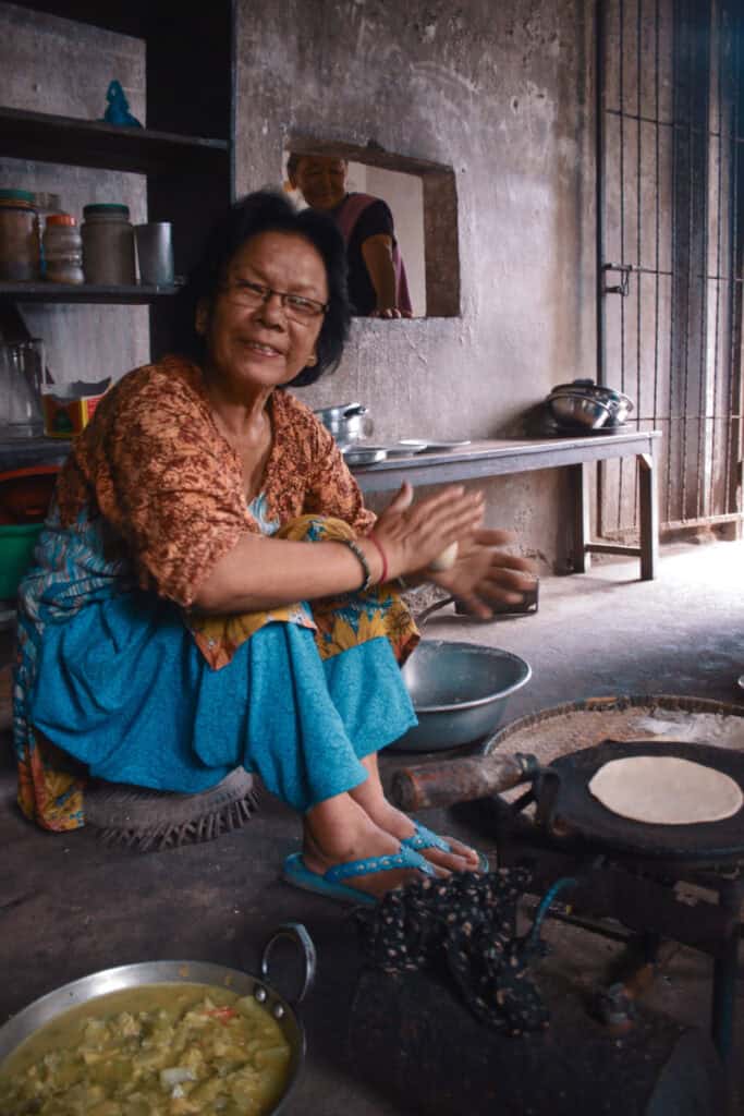 Ältere nepalesische Frau beim Zubereiten von Dal Bhat, lachend in einer traditionellen Küche in Nepal.