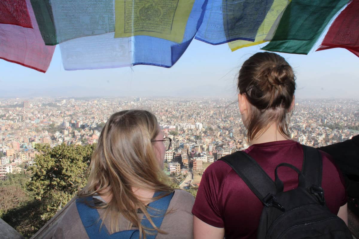 Zwei Reisende blicken unter bunten Gebetsfahnen über die Dächer von Kathmandu