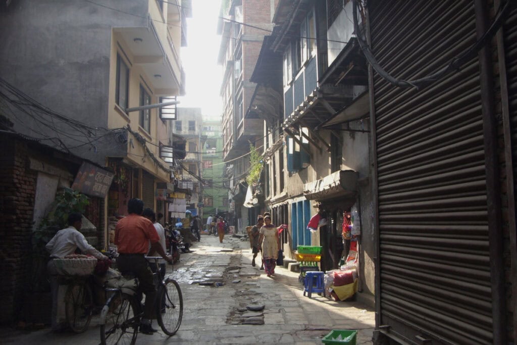 Schmale Gasse in einem nepalesischen Wohnviertel von Kathmandu mit Fahrrädern, kleinen Läden und Bewohnern am frühen Morgen.