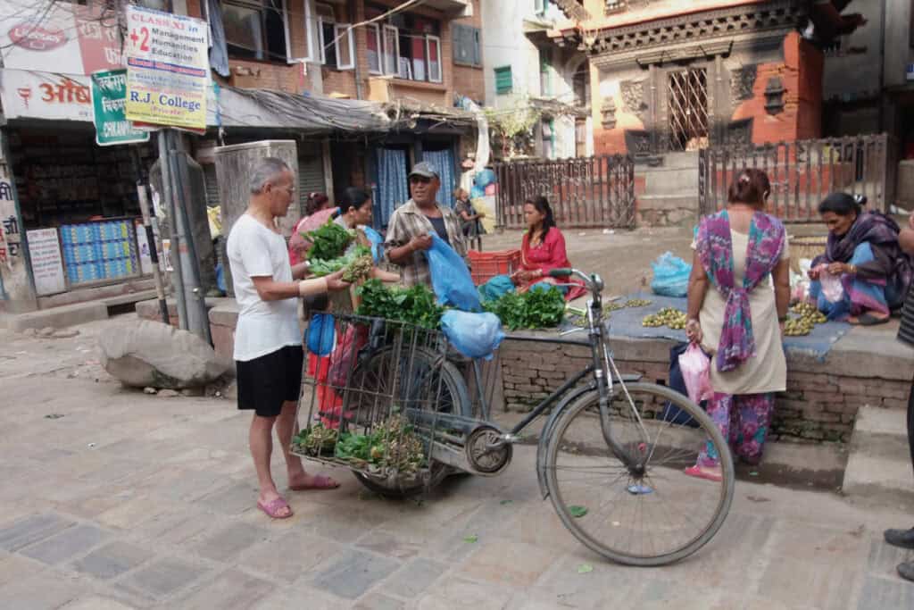 Menschen kaufen frisches Gemüse bei einem Händler mit Fahrrad auf einem kleinen Markt in Kathmandu – typische Alltagsszene in Nepal.