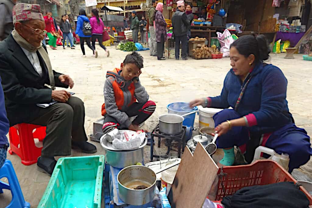Eine Frau bereitet in einer Gasse in Kathmandu heißen Tee zu, während ein älterer Mann und ein Junge warten – typische Alltagsszene in Nepal.