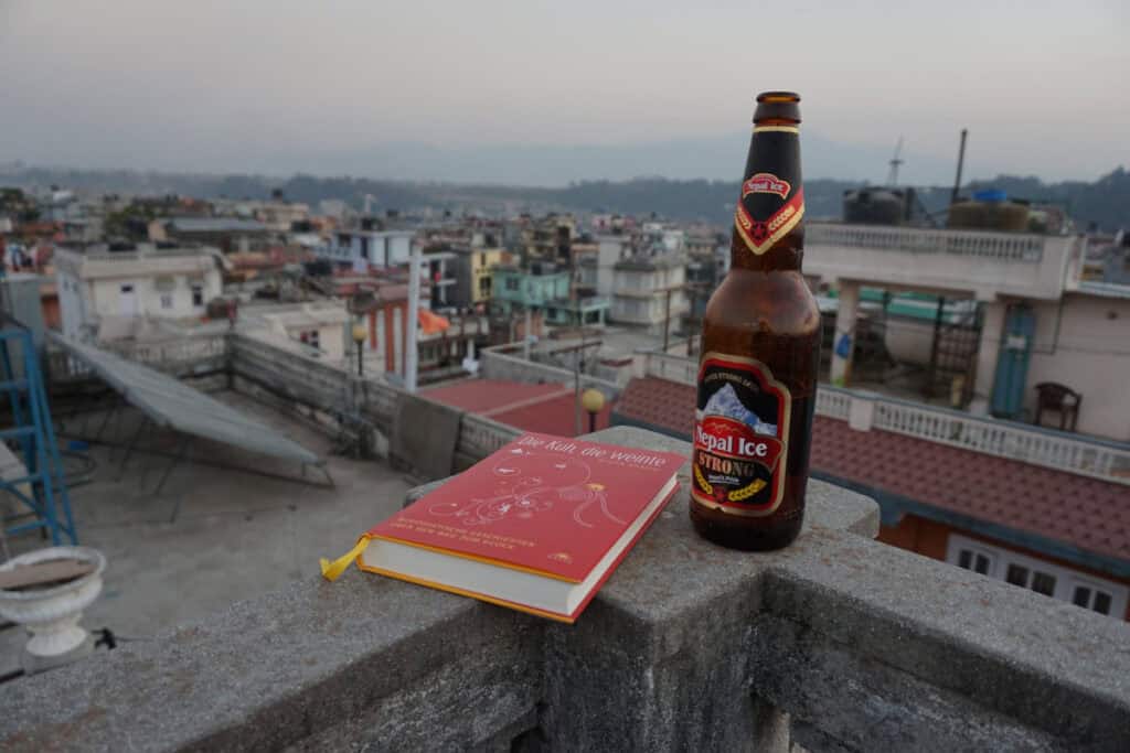 Eine Flasche Nepal Ice Bier und das Buch „Die Kuh, die weinte“ auf einer Dachterrasse mit Blick über Kathmandu.