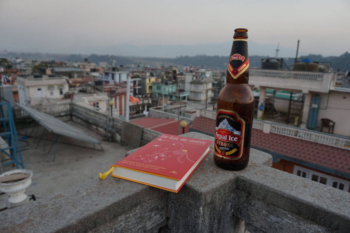 Eine Flasche Nepal Ice Bier und das Buch „Die Kuh, die weinte“ auf einer Dachterrasse mit Blick über Kathmandu.