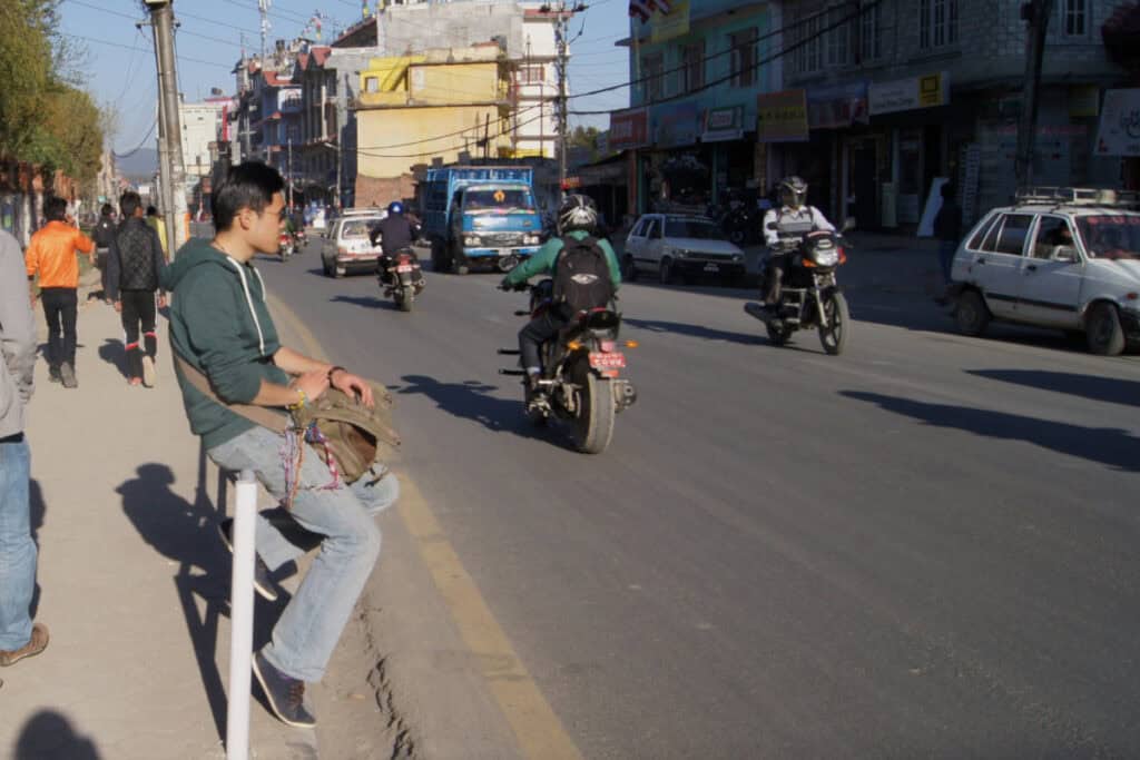 Ein Mann sitzt gelassen am Straßenrand in Kathmandu und wartet auf den Bus – ein alltäglicher Moment, der die nepalesische Gelassenheit im Umgang mit Zeit widerspiegelt.