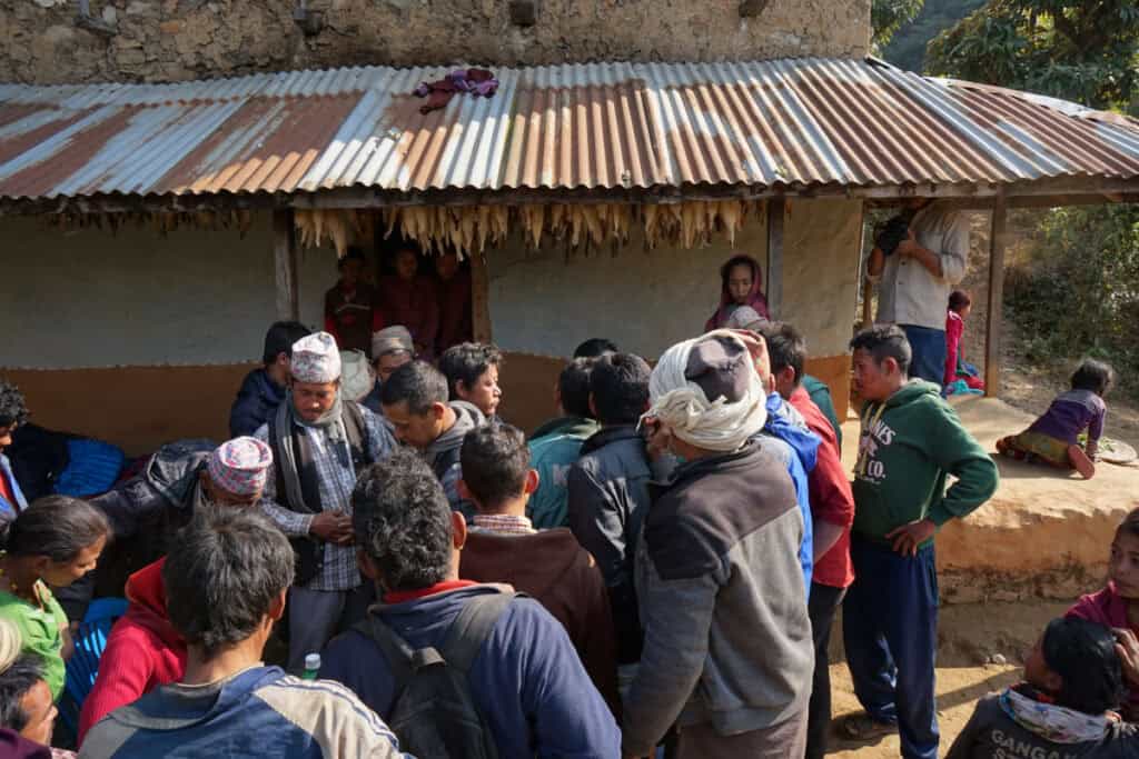 Dorfversammlung in Ikudol: Zahlreiche Männer diskutieren lautstark vor einem einfachen Haus, während Kinder und Zuschauer am Rand sitzen.