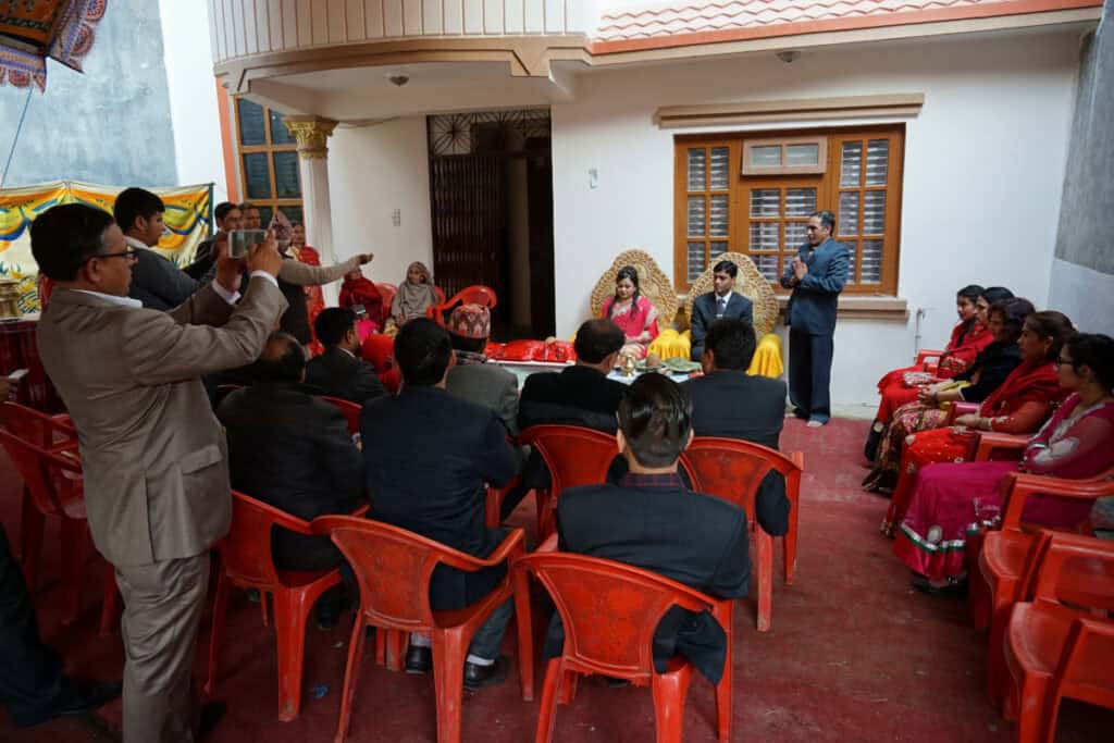 Nepalesische Hochzeitsgesellschaft mit klarer Sitzordnung: Männer sitzen dem Brautpaar zugewandt, Frauen am Rand.