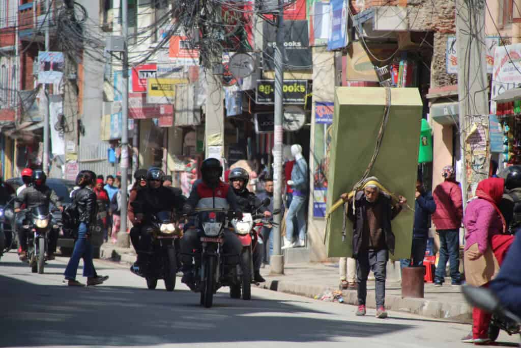 Mann trägt schwere Last durch eine belebte Straße in Nepal – informelle Arbeit im städtischen Alltag.
