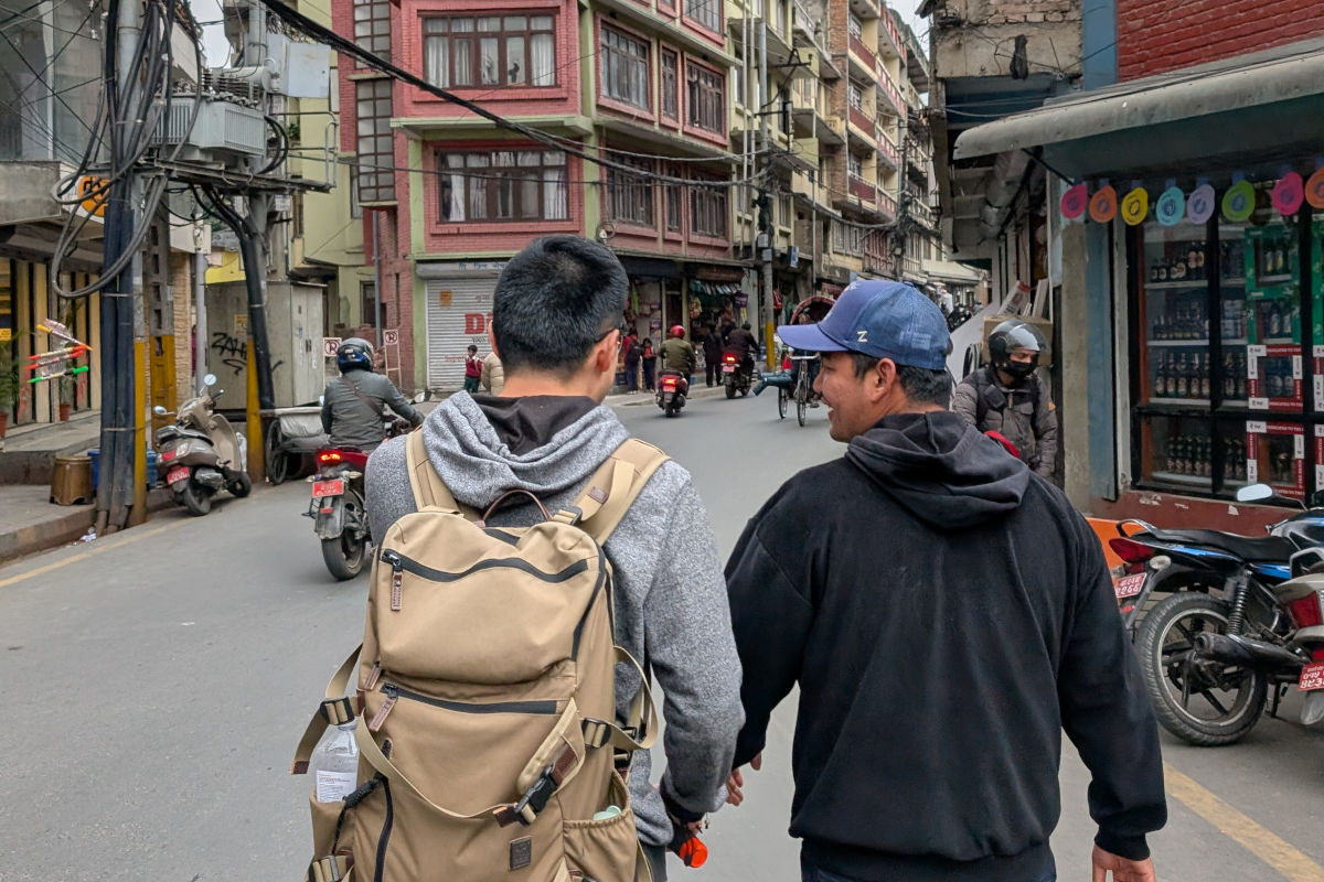 Zwei Männer gehen im Gespräch durch eine Straße in Kathmandu mit Motorrädern und Häusern im Hintergrund