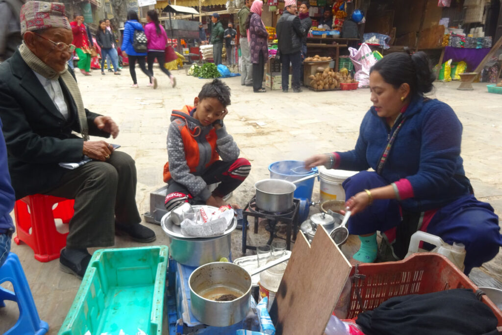 Frau kocht auf der Straße in Kathmandu während ein Kind und ein älterer Mann daneben sitzen